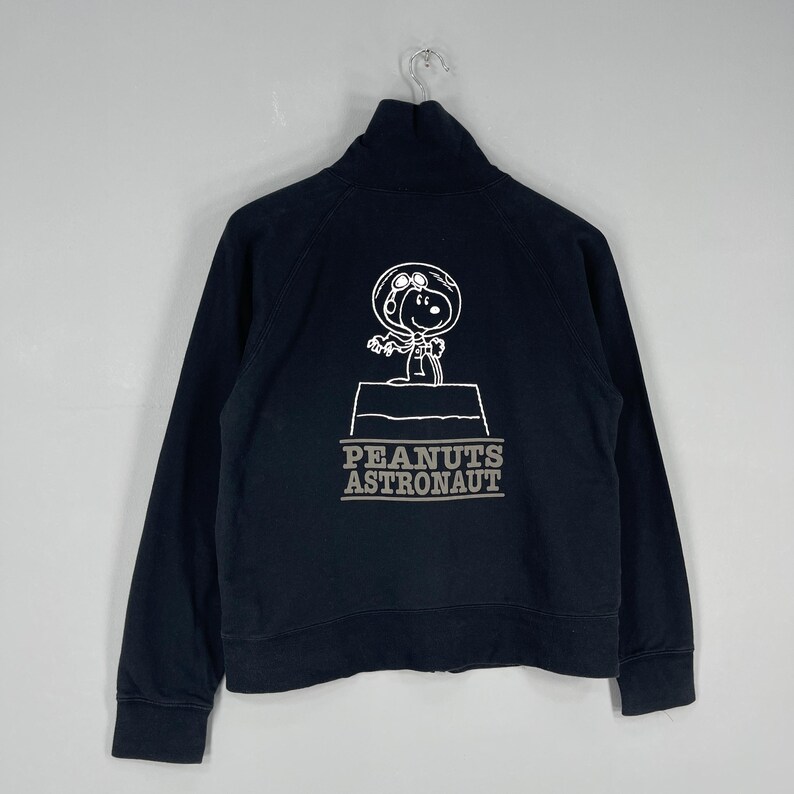 May include: Black zip-up sweatshirt featuring a white graphic of Snoopy in an astronaut helmet, standing on a platform. The text "PEANUTS ASTRONAUT" is below the graphic. The sweatshirt has a high collar and long sleeves.