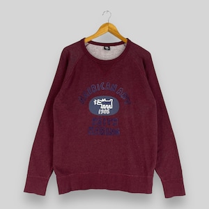Puede incluir: Una sudadera color burdeos con un gr&aacute;fico azul y blanco de un perro y el texto "American Art 1985 Keith Haring" en la parte delantera.