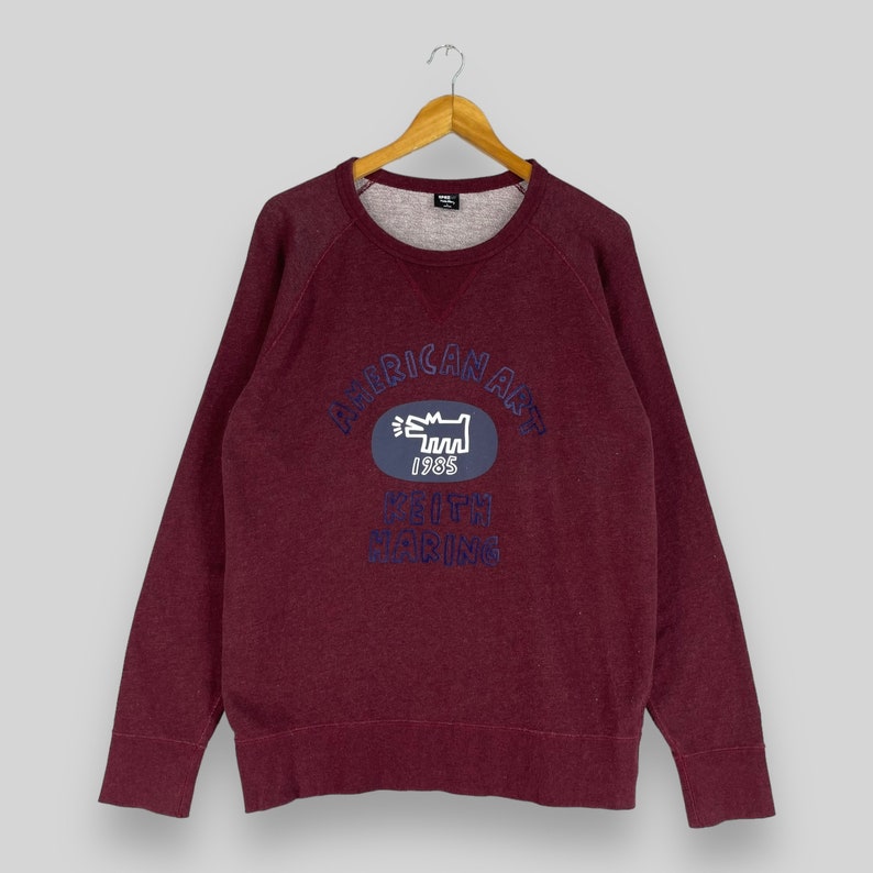 Puede incluir: Una sudadera color burdeos con un gr&aacute;fico azul y blanco de un perro y el texto "American Art 1985 Keith Haring" en la parte delantera.