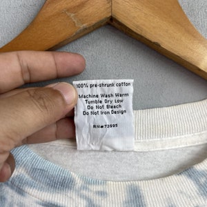 May include: White cotton t-shirt with a blue and white tie-dye pattern. The care label says "100% pre-shrunk cotton, Machine Wash Warm, Tumble Dry Low, Do Not Bleach, Do Not Iron Design, RN#73995".