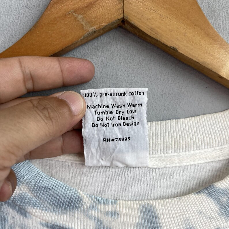 May include: White cotton t-shirt with a blue and white tie-dye pattern. The care label says "100% pre-shrunk cotton, Machine Wash Warm, Tumble Dry Low, Do Not Bleach, Do Not Iron Design, RN#73995".