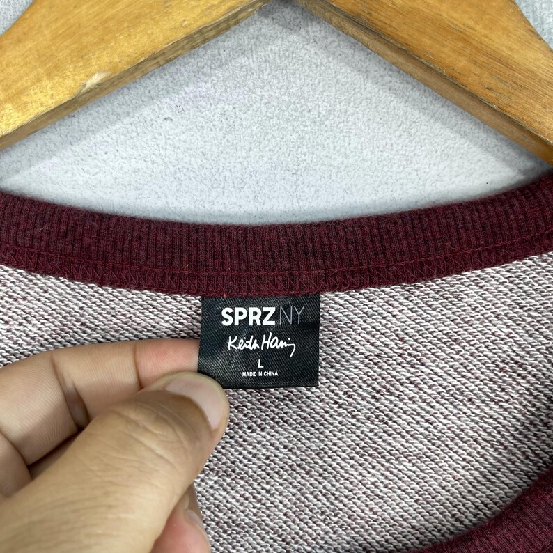 Puede incluir: Una sudadera color burdeos con una etiqueta negra que dice "SPRZNY Keda Hari" y "L" para el tama&ntilde;o. La etiqueta tambi&eacute;n dice "Hecho en China".