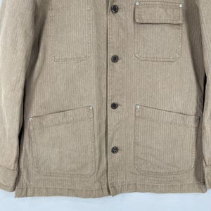 May include: A tan corduroy jacket with four front pockets and a chest pocket. The jacket has a button-up closure and a classic collar. The fabric has a vertical ribbed texture. The jacket is a neutral color and suitable for various outfits.