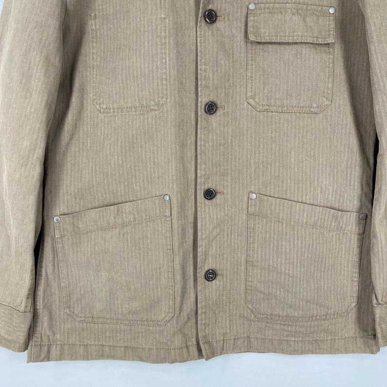 May include: A tan corduroy jacket with four front pockets and a chest pocket. The jacket has a button-up closure and a classic collar. The fabric has a vertical ribbed texture. The jacket is a neutral color and suitable for various outfits.