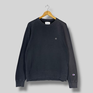 Puede incluir: Sudadera Champion gris oscuro con un pequeño logotipo en el pecho y un logotipo más grande en el puño izquierdo. La sudadera está colgada de una percha de madera. La tela parece desgastada y tiene una estética vintage.