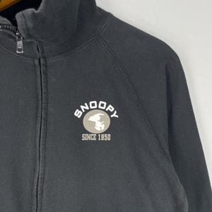 May include: Black zip-up sweatshirt featuring a Snoopy graphic on the chest. The graphic includes the word "Snoopy", an image of Snoopy, and the text "Since 1950". The sweatshirt has a full zipper and a high collar.