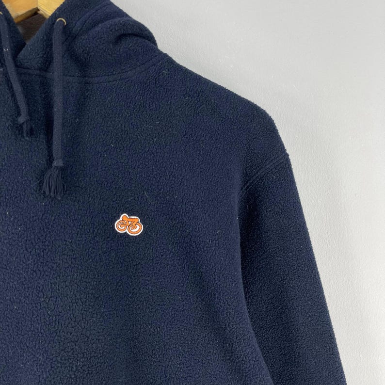 Puede incluir: Sudadera con capucha de forro polar azul marino con capucha con cord&oacute;n. La sudadera tiene un peque&ntilde;o emblema naranja y marr&oacute;n en el pecho. La tela tiene un aspecto suave y texturizado. Es una prenda informal.