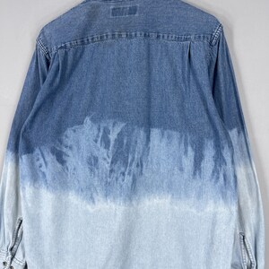 May include: A denim shirt with a unique, faded blue and white tie-dye effect. The long-sleeved shirt features a classic collar, button-down front, and buttoned cuffs. The back of the shirt is visible, showcasing the dye pattern.
