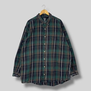 Puede incluir: Una camisa de franela a cuadros con botones y mangas largas en tonos verdes, azul marino, marrón y blanco. La camisa está colgada de una percha de madera. La camisa tiene cuello abotonado y un bolsillo en el pecho.