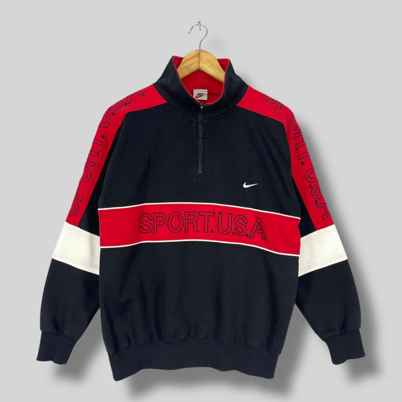 Może przedstawiać: Czarna, czerwona i biała bluza z zamkiem błyskawicznym na &frac14; długości z napisem "SPORT.U.S.A." z przodu i logo Nike swoosh na prawym rękawie.