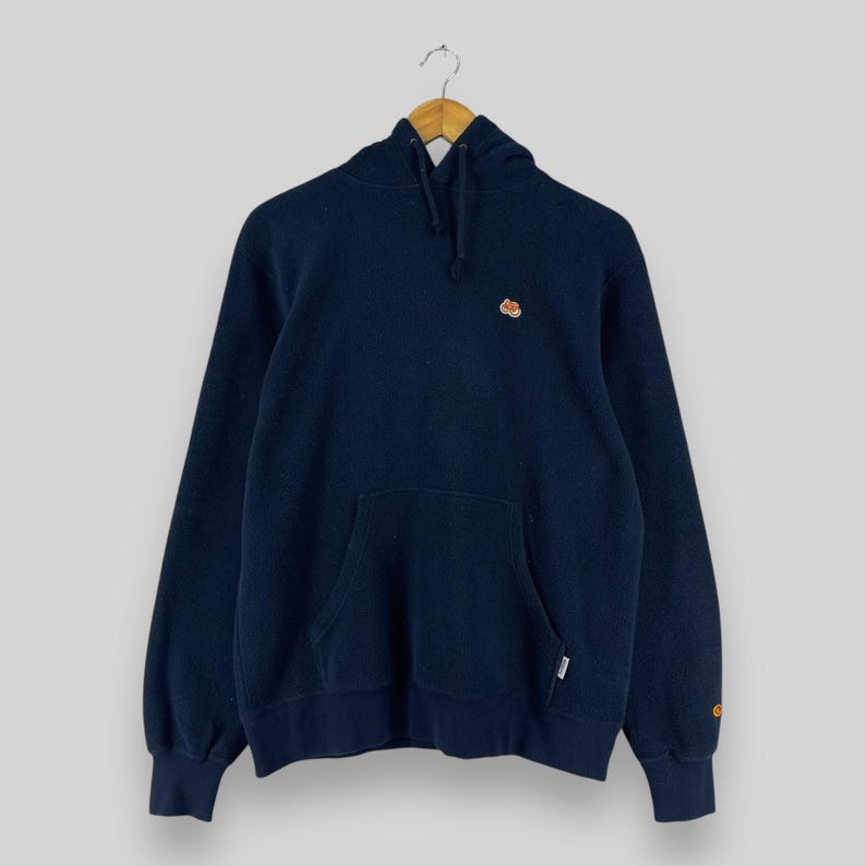 Puede incluir: Sudadera con capucha azul marino con un bolsillo delantero y un peque&ntilde;o dise&ntilde;o bordado en el pecho. La sudadera tiene una capucha con cord&oacute;n y pu&ntilde;os acanalados. La prenda est&aacute; colgada en una percha de madera.