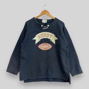 Puede incluir: Una sudadera negra de manga larga con un diseño de temática de rugby. La sudadera presenta una pancarta blanca con la palabra "RUGBY" en letras rojas, una pelota de fútbol marrón y blanca, y el texto "TROPICAL CLUB".