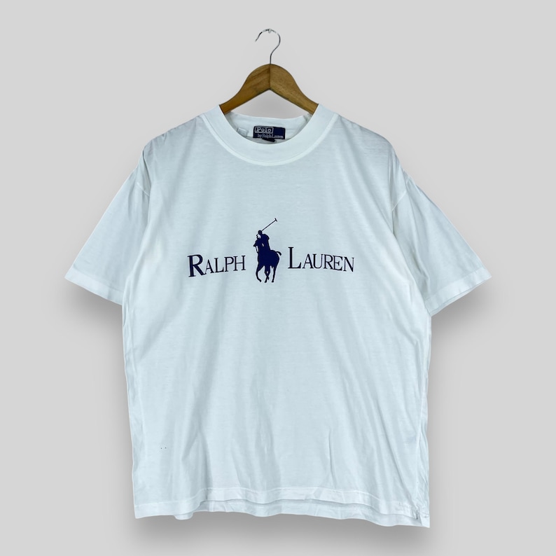 Puede incluir: Camiseta blanca de manga corta con un logotipo de jugador de polo azul marino y el texto "RALPH LAUREN" impreso en la parte delantera.