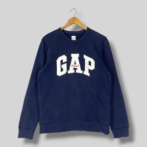 Peut inclure: Un sweat-shirt bleu marine à manches longues arborant un grand logo GAP blanc en arc de cercle sur la poitrine, avec le mot Original écrit en petites lettres rouges en dessous. Le vêtement possède un col rond et est sur un cintre en bois.