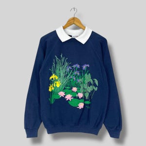 Puede incluir: Sudadera azul marino con cuello blanco, con una escena de estanque colorida. El diseño incluye lirios amarillos y morados, nenúfares rosas y follaje verde. La sudadera está colgada en una percha de madera.