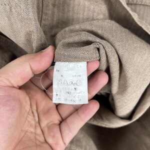 May include: Close-up of a tan fabric with a herringbone pattern. A white tag with Japanese text and symbols is visible. The fabric is held by a hand.
