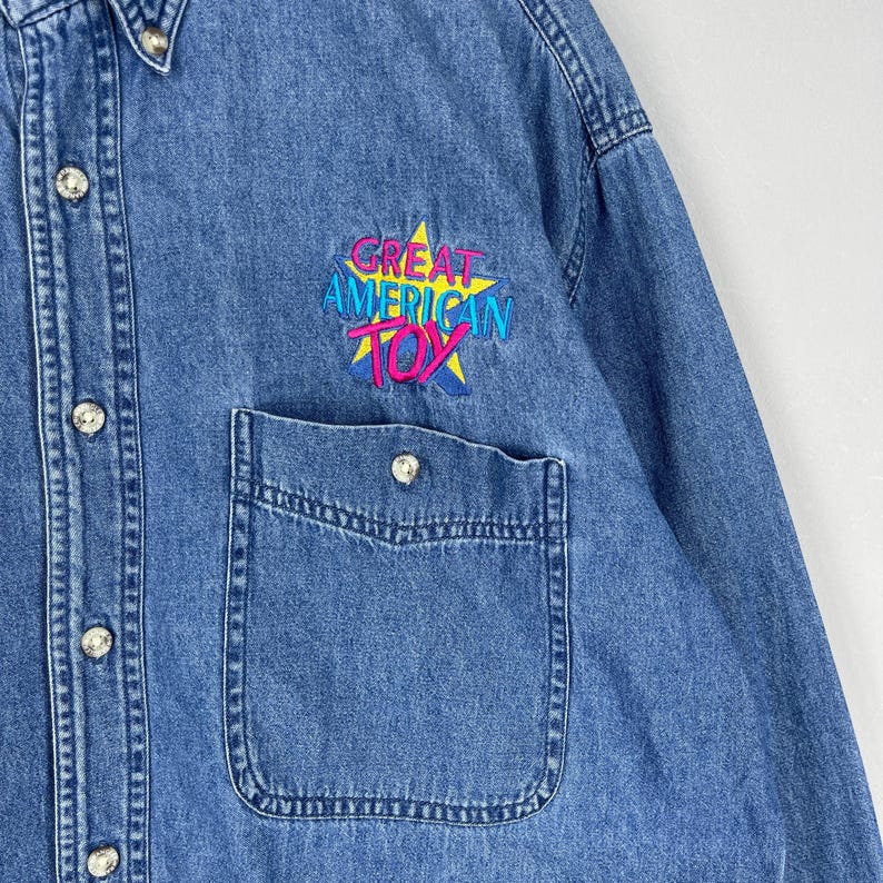 Op de afbeelding: Een blauw spijkeroverhemd met knopen en een borstzak. De tekst "GREAT AMERICAN TOY" is in kleurrijk borduursel aangebracht. Het overhemd heeft zilveren knopen en een klassieke kraag.