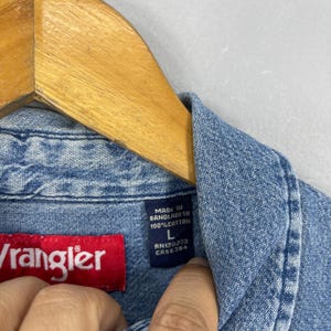 May include: Close-up of a light blue denim Wrangler shirt. The shirt has a red Wrangler label and a blue label with text that reads "MADE IN BANGLADESH 100% COTTON L RN130273 CA55394". The shirt is hanging on a wooden hanger.
