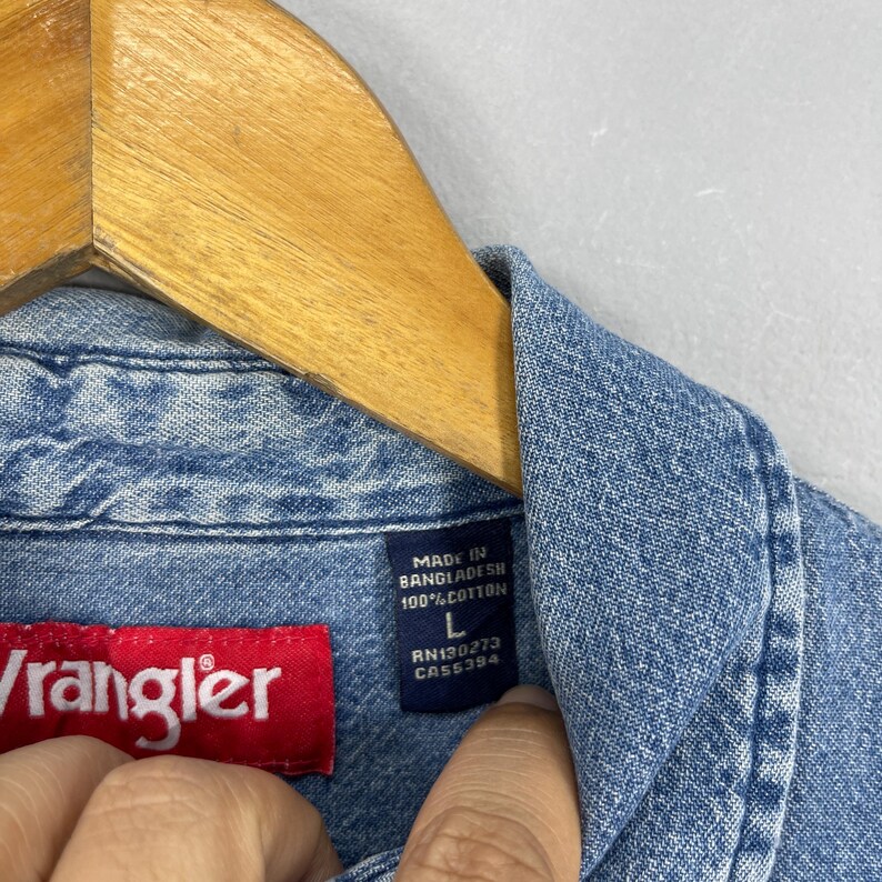 May include: Close-up of a light blue denim Wrangler shirt. The shirt has a red Wrangler label and a blue label with text that reads "MADE IN BANGLADESH 100% COTTON L RN130273 CA55394". The shirt is hanging on a wooden hanger.