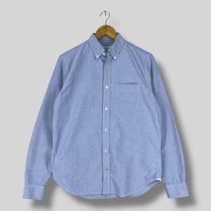 Könnte beinhalten: Hellblaues Hemd mit langen Ärmeln, Button-Down-Kragen und Brusttasche. Das Hemd hängt an einem Holzbügel vor einem hellgrauen Hintergrund. Der Stoff hat eine feine Textur.