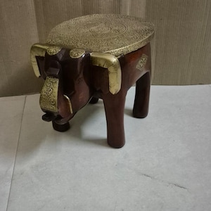 May include: A wooden elephant stool with a round, brass top. The top has an intricate, engraved design. The elephant's ears and tusks are also made of brass.