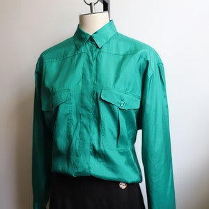 May include: A teal blue long-sleeved button-up shirt with two large pockets on the front. The shirt is on a mannequin and paired with a black skirt.