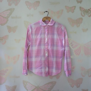 Pode incluir: Uma camisa rosa xadrez de manga comprida com gola franzida, frente abotoada e punhos abotoados. A camisa está pendurada em um cabide de madeira contra um fundo de papel de parede com padrões de borboletas e libélulas.