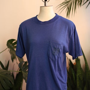 May include: A blue t-shirt with a small pocket on the left side. The pocket has a small embroidered design.