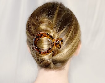 May include: A brown and tortoiseshell hair claw with a large oval opening. The claw is holding a woman's blonde hair in a bun.