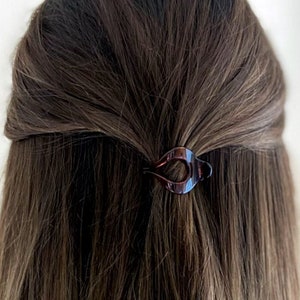 May include: A brown tortoiseshell hair clip securing a section of long, wavy brown hair. The clip is a small, decorative accessory with a unique shape, adding a touch of style to the hairstyle. The hair is styled in a half-up, half-down fashion.