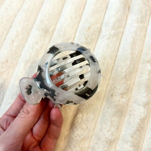 May include: A white and black tortoise shell hair claw with a comb design. The claw is round and has a smooth finish.