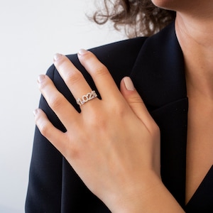 Può includere: Un anello d'argento con il numero 2022 in un carattere stilizzato. L'anello è incastonato con piccole pietre trasparenti.