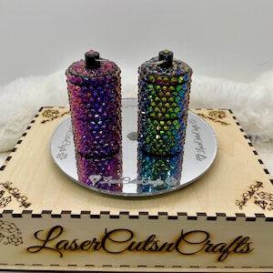 May include: Two small, cylindrical containers covered in iridescent rhinestones. The containers are on a mirrored surface, which is on a wooden box with the text "Laser Cuts n Crafts" engraved on the front.