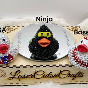 Puede incluir: Tres patos de goma decorados con pedrer&iacute;a. El pato de la izquierda es blanco con el texto USA en rojo y azul. El pato central es negro con una m&aacute;scara amarilla y pico naranja, etiquetado como Ninja. El pato de la derecha es blanco con costuras de b&eacute;isbol rojas.