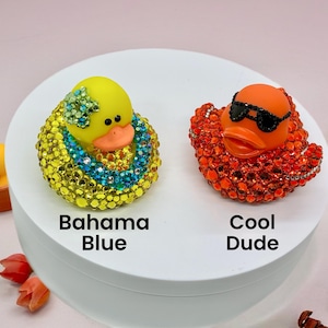 Puede incluir: Dos patos de goma decorados con pedrer&iacute;a. El pato de la izquierda es amarillo con un collar y adorno de cabeza de pedrer&iacute;a azul y verde, etiquetado Bahama Blue. El pato de la derecha es naranja con gafas de sol de pedrer&iacute;a negra, etiquetado Cool Dude.