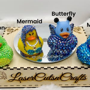 Puede incluir: Cuatro patos de goma decorados con pedrer&iacute;a, etiquetados como Dragon, Mermaid, Butterfly y Yellow Mohawk. Los patos se exhiben sobre un espejo circular encima de una caja de madera con el texto LaserCutsnCrafts y Bazaart impreso al frente.