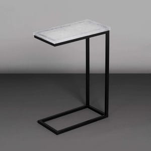 Könnte beinhalten: Ein moderner Beistelltisch mit einer rechteckigen, hellfarbigen Platte und einem schwarzen Metallrahmen. Das Design des Tisches zeichnet sich durch eine auskragende Platte aus, die von einem vertikalen Bein und einem Sockel getragen wird, der sich entlang des Bodens erstreckt. Der Tisch ist etwa 61 cm lang.