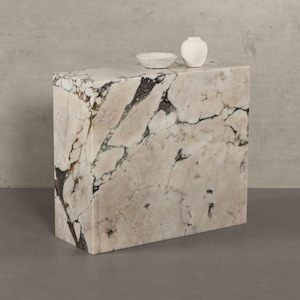 Pode incluir: Uma mesa lateral retangular de mármore com um padrão de veias creme e cinza. Um pequeno vaso branco e uma tigela salpicada estão no topo. As dimensões da mesa não são visíveis na imagem.