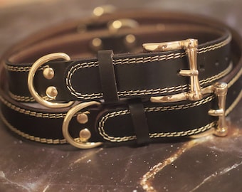 Puede incluir: Dos collares de perro de cuero negro con herrajes dorados. Los collares están cosidos con un hilo dorado fino. Los collares están apilados uno encima del otro.