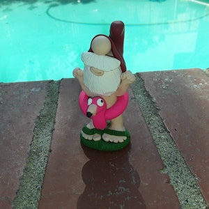 May include: A small, white bearded gnome figurine with a red hat and a pink flamingo pool float. The gnome is standing on a green base and is holding the flamingo float in front of him. The gnome is standing on a brick surface.