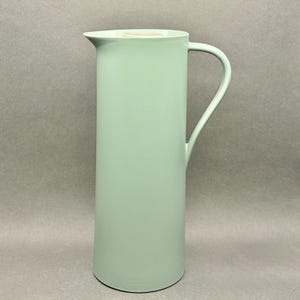 May include: A tall, pale green jug with a curved handle and a spout. The jug has a cylindrical body and a slightly rounded base. The handle is thin and curves gracefully from the top to the side of the jug. The jug is made of ceramic or a similar material.