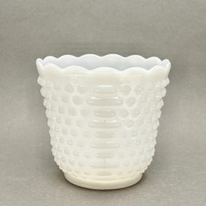 Könnte beinhalten: Eine weiße Hobnail-Glasvase mit gewelltem Rand. Die Vase hat eine strukturierte Oberfläche mit kleinen, erhabenen Noppen und horizontalen Bändern. Die Gesamtform ist zylindrisch, mit einem leicht ausgestellten oberen Rand und einem abgerundeten Boden.