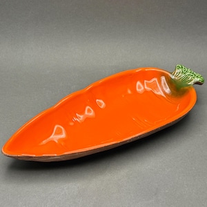 Puede incluir: Plato de cerámica naranja con forma de zanahoria. El plato tiene un interior naranja brillante y un borde marrón. El tallo es verde con un diseño de hojas texturizadas. Una pieza decorativa para servir aperitivos o entremeses.