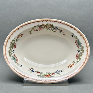 May include: Oval-shaped ceramic serving dish with a cream-colored interior and a decorative floral border. The rim features a scalloped design in a reddish-brown hue, with colorful floral and foliage accents. The dish is displayed on a clear stand.