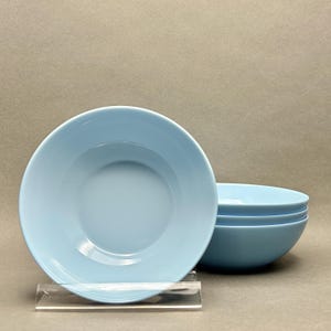 May include: A set of four light blue ceramic bowls. The bowls are round with a slightly flared rim and a smooth, glossy finish. One bowl is in the foreground, and three are stacked behind it. The bowls are displayed on a clear acrylic stand.