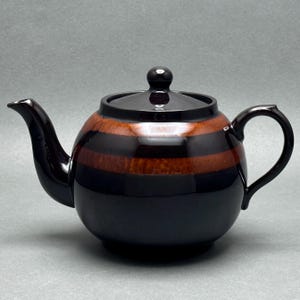 May include: A ceramic teapot with a glossy finish. The teapot is primarily black with two horizontal bands of brown and a matching lid. It has a curved spout and a rounded handle. The teapot is designed for brewing tea.