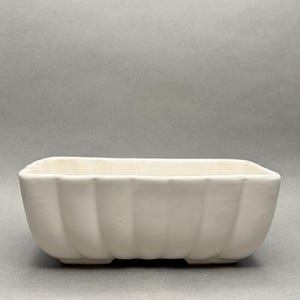 May include: A rectangular white ceramic planter with a scalloped, fluted exterior design. The vessel features a smooth, matte finish and a wide, open top, resting on a slightly elevated base with visible vertical ridges along all four sides.