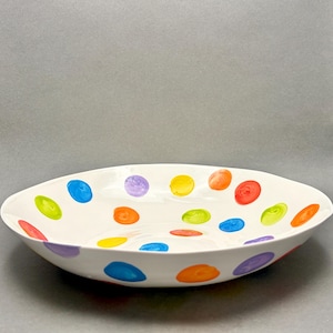 Può includere: Una ciotola in ceramica bianca dalla forma irregolare e organica, caratterizzata da pois dipinti a mano e sparsi in tonalità vivaci di blu, giallo, arancione, rosso, verde e viola sulle superfici interne ed esterne.