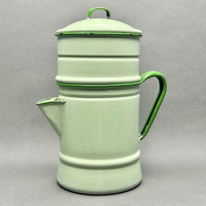 Puede incluir: Una cafetera de esmalte en color verde menta claro con tapa a juego y asa de color verde oscuro. El recipiente cilíndrico cuenta con un pico lateral y un diseño escalonado con bordes en verde oscuro que muestran un ligero desgaste.