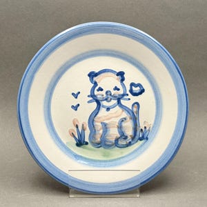 Könnte beinhalten: Ein weißer Keramikteller mit blauem Rand und einer skurrilen Katzenillustration. Die Katze ist in Blau und Rosa dargestellt, mit einer blauen Kontur und einer kleinen blauen Wolke mit einem Kuss. Der Teller hat einen Durchmesser von etwa 20 cm.